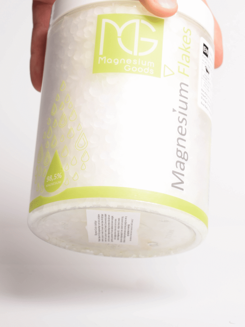 Hand hält Behälter mit Magnesium Flakes von Magnesium Goods, weiße Flocken und grünes Etikett