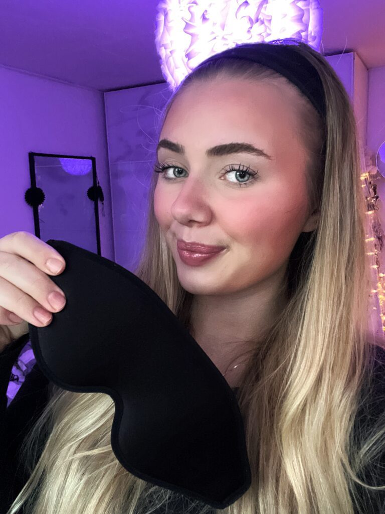 Selfie einer jungen Frau mit langem blondem Haar und Stirnband, die in einem lila beleuchteten Zimmer eine schwarze Schlafmaske hält