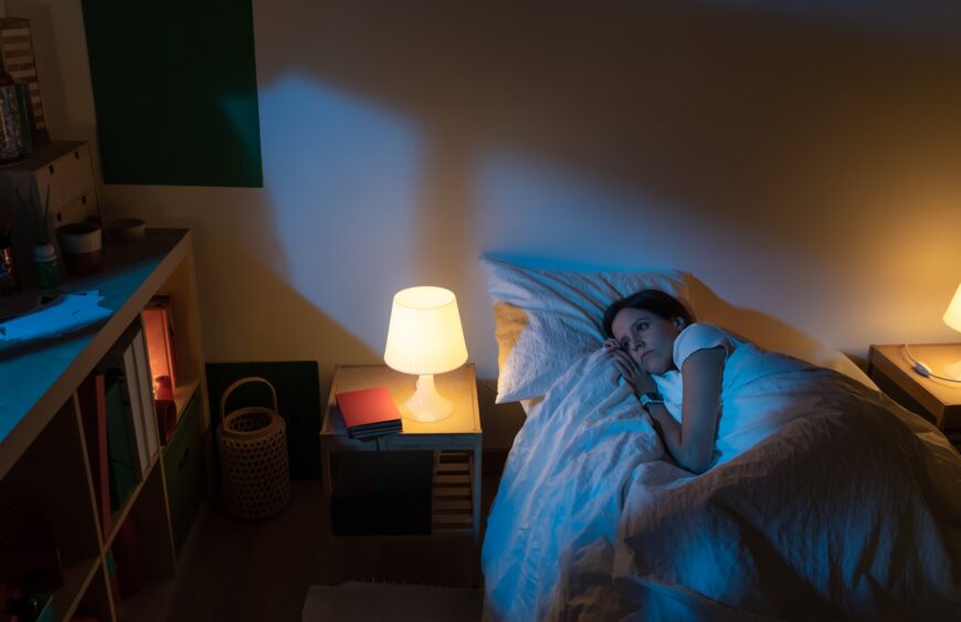 Junge Frau mit Schlaflosigkeit liegt wach im Bett neben einem Nachttisch mit eingeschalteter Lampe