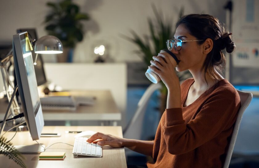 Junge Frau mit Brille trinkt Coffee to go und arbeitet abends im Büro am Computer