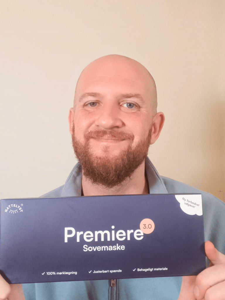 Lächelnder Mann mit Bart hält die Premiere 3.0 Schlafmaske in dunkelblauer Verpackung