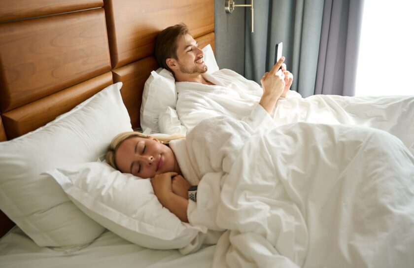 Mann im Bademantel liegt im großen Bett und schaut aufs Handy, daneben schläft eine Frau unter weißer Bettdecke