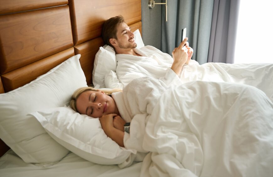 Mann im Bademantel liegt im großen Bett und schaut aufs Handy, daneben schläft eine Frau unter weißer Bettdecke