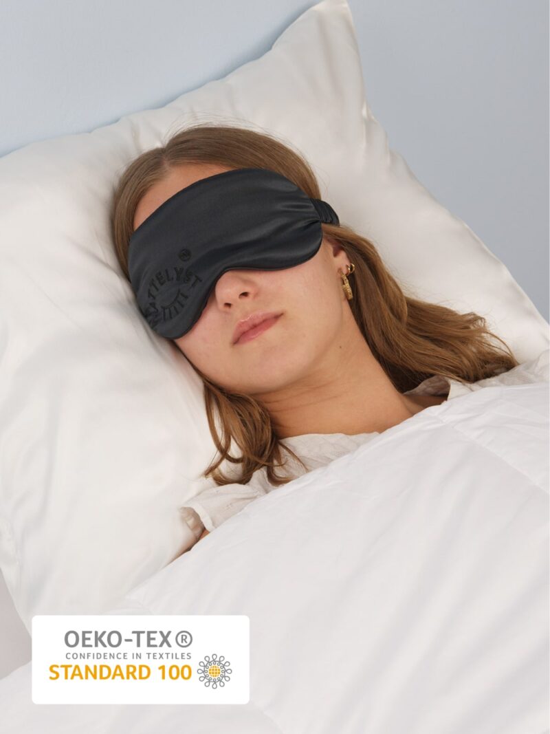 Ung kvinde sover med sort silkeøjenmaske på hvid pude og dyne, OEKO-TEX Standard 100 mærke