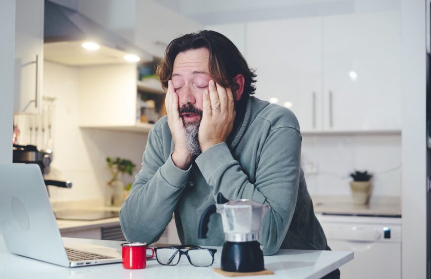 Morgenmüder Mann in der Küche stützt das Gesicht in die Hände, daneben Laptop, Kaffeekanne, rote Tasse und Brille