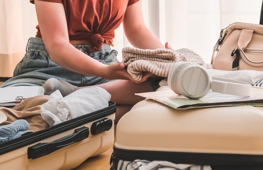 Frau packt einen Koffer mit gefalteter Kleidung und Kopfhörern, daneben beige Koffer und Reisetasche