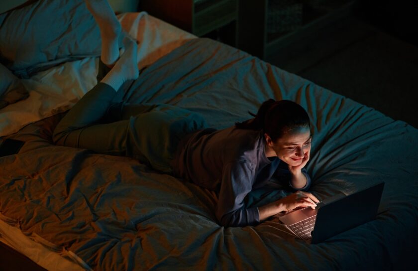 Junge Frau liegt nachts im Bett und lächelt im blauen Licht ihres Laptops, während sie einen Film streamt