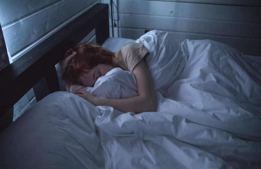 Person mit roten Haaren schläft in eine weiße Bettdecke gehüllt in einem dunklen Holzbett bei gedämpftem blauem Licht
