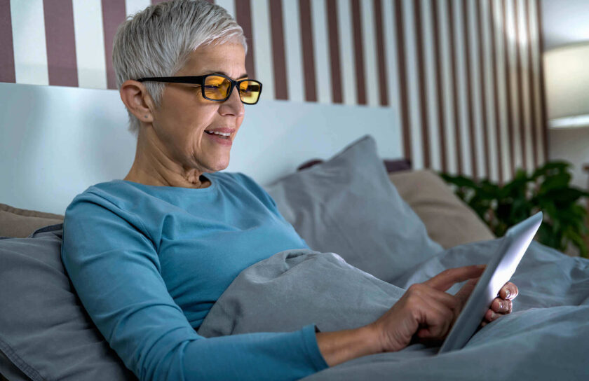 Frau im Bett mit Blaulichtbrille schaut auf ihr Smartphone