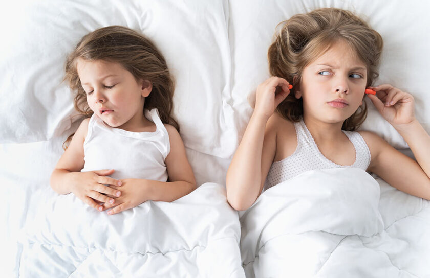 Zwei Mädchen im Bett: eines schläft, das andere setzt genervt Ohrstöpsel ein