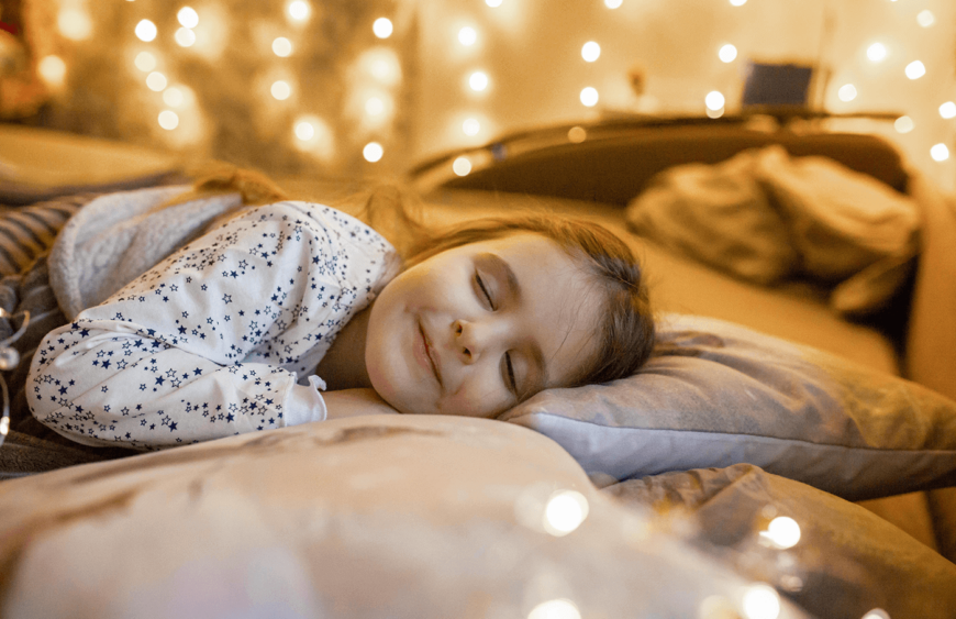 Schlafendes Mädchen im Sternen-Pyjama auf einem Kissen, umgeben von warmen Lichterketten