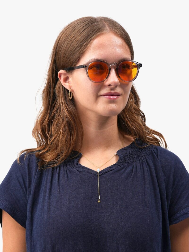 Frau mit braunem Haar, runder orangefarbener Brille, dunkelblauer Bluse, goldenem Anhänger und kleinen goldenen Creolen