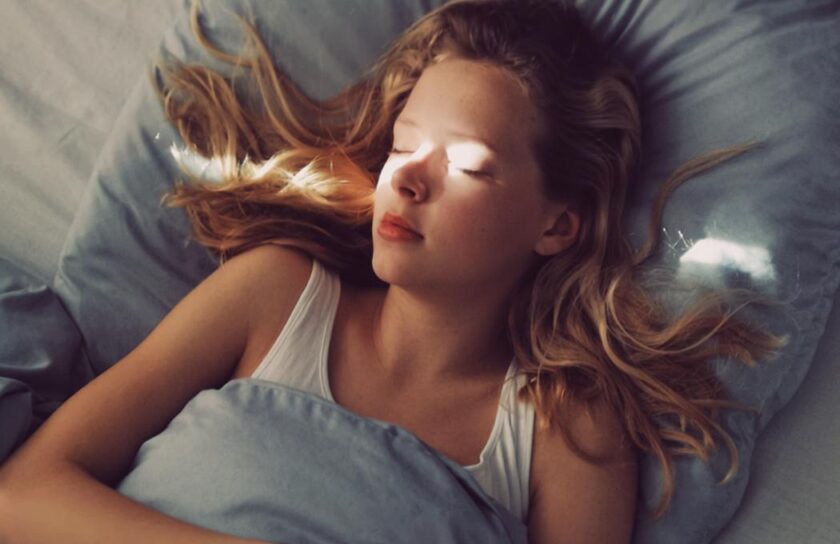 Junge Frau schläft, das erste Morgenlicht fällt auf ihr Gesicht unter blauer Bettwäsche