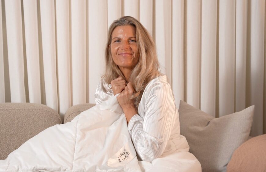Lächelnde Frau im weißen Pyjama hält eine weiche Bettdecke auf dem Sofa vor beigen Vorhängen