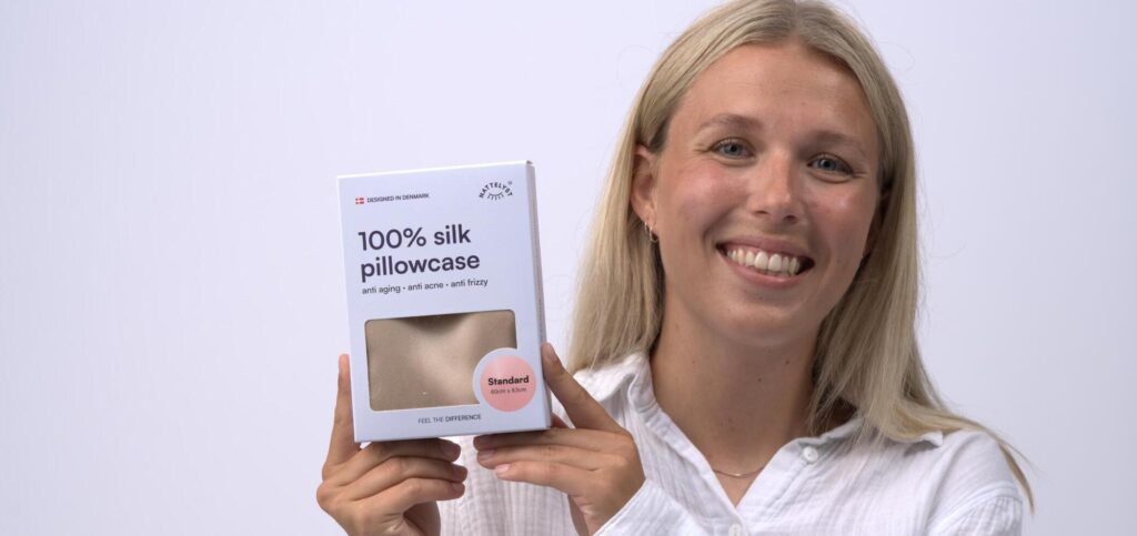 Lächelnde Frau hält eine Verpackung mit Seidenkissenbezug aus 100 % Seide in Beige, Standardgröße