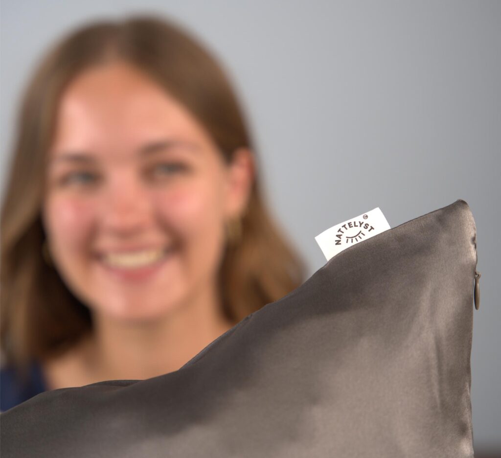 Lächelnde Frau hält einen grauen Seidenkissenbezug mit sichtbarem Nattelyst Label