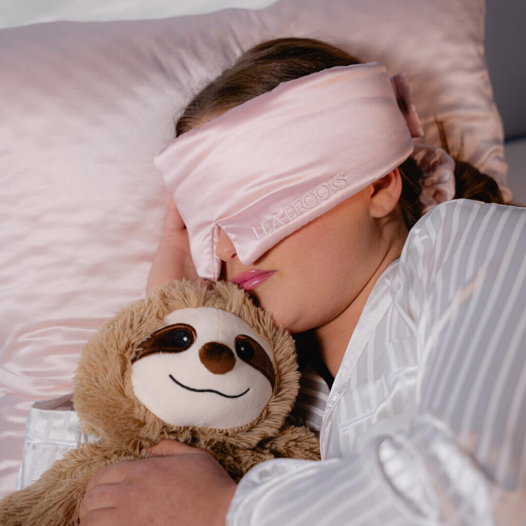 Person mit rosafarbener Schlafmaske und gestreiftem Pyjama kuschelt mit einem weichen Faultier-Kuscheltier auf einem rosafarbenen Seidenkissen