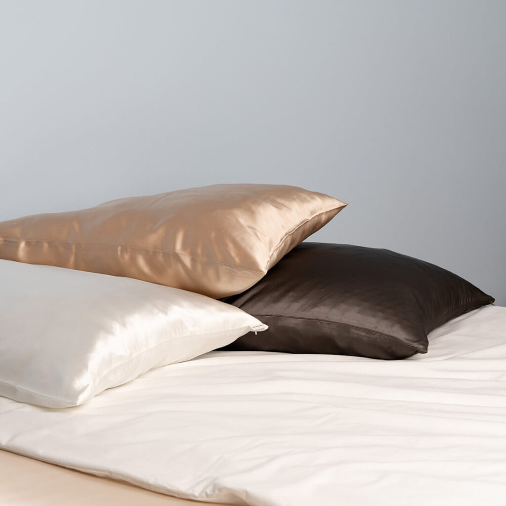 Drei luxuriöse Satinkissen in Gold, Braun und Creme auf weißem Laken