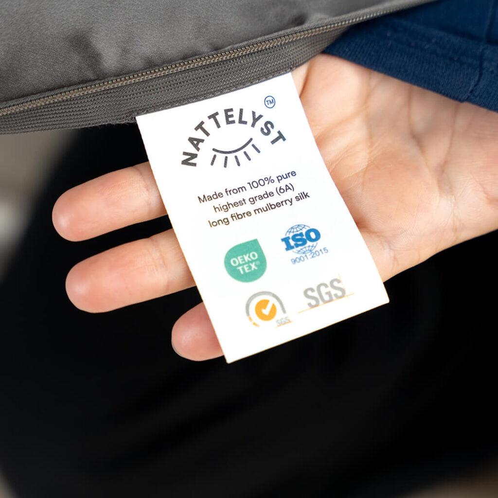 Hand hält weißes Label mit NATTELYST Logo, Hinweis auf 100 % Maulbeerseide sowie OEKO-TEX-, ISO- und SGS-Zertifikate
