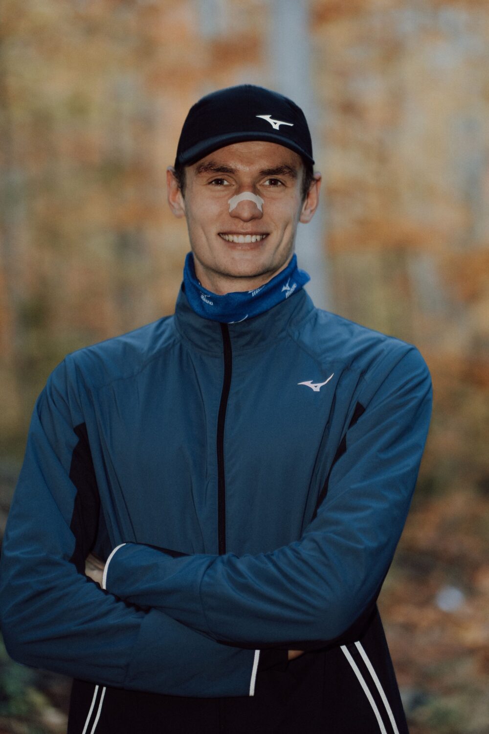 Lächelnder Läufer in blauer Jacke und Cap mit Nasenpflaster, Arme verschränkt im Herbstwald
