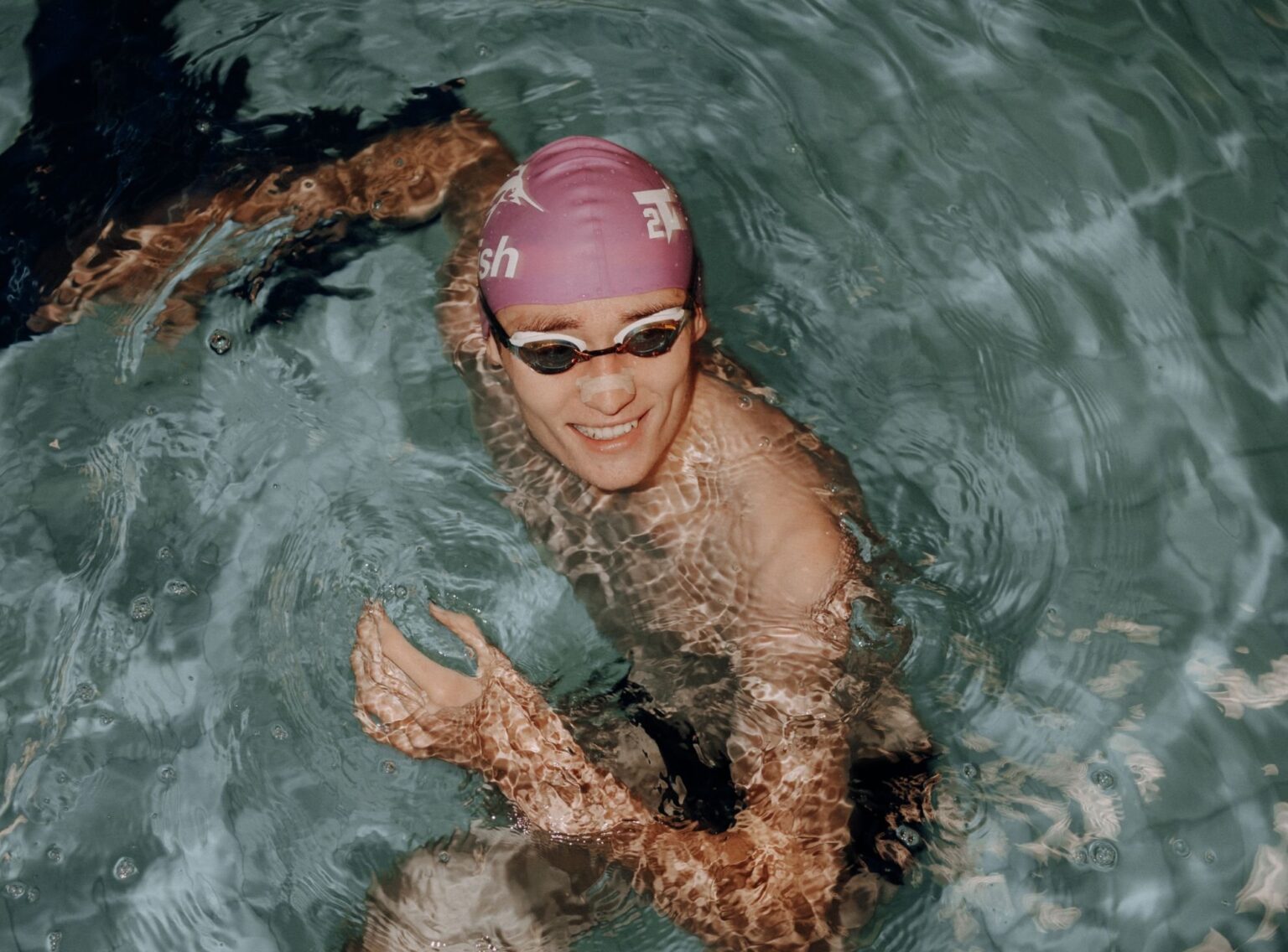 Lächelnder Schwimmer mit lila Badekappe und Schwimmbrille im Pool, Wasserreflexe auf Arm und Schultern