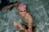 Lächelnder Schwimmer mit lila Badekappe und Schwimmbrille im Pool, Wasserreflexe auf Arm und Schultern