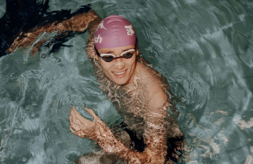 Lächelnder Schwimmer mit lila Badekappe und Schwimmbrille im Pool, Wasserreflexe auf Arm und Schultern