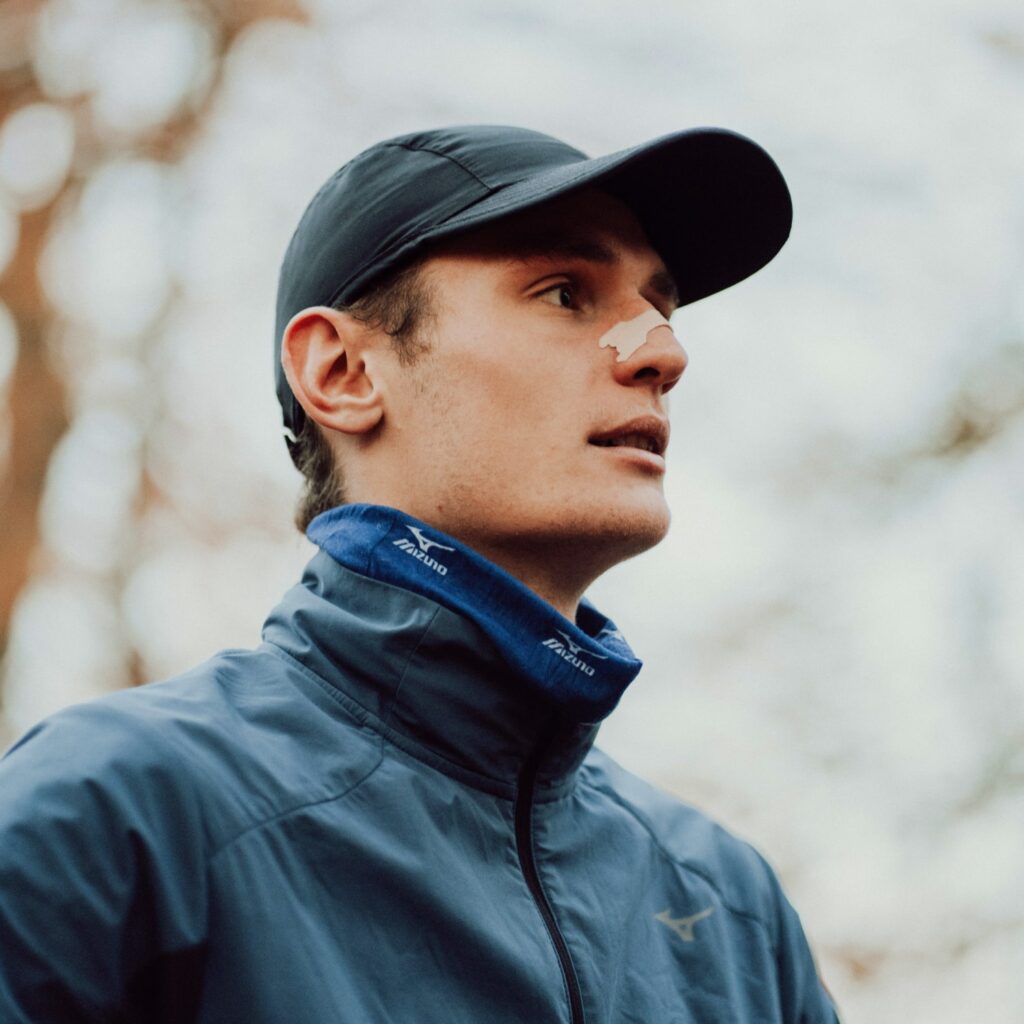 Läufer in blauer Laufjacke und Cap mit Nasenpflaster im Wald, Blick zur Seite