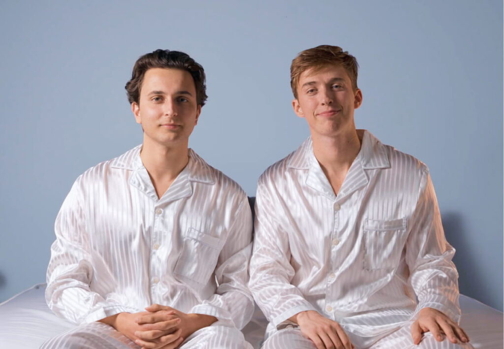 Zwei junge Männer in passenden weißen Satinpyjamas sitzen vor einer blauen Wand auf dem Bett
