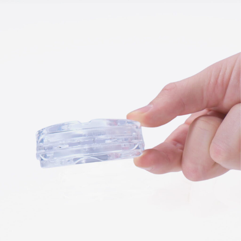 Hand hält eine kleine runde, transparente Kunststoffdose mit Innenfächern und Rille vor weißem Hintergrund