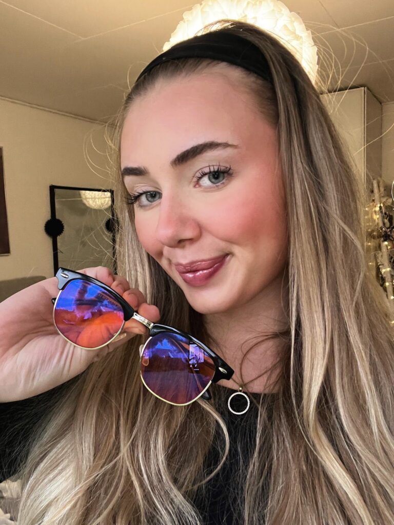 Porträt einer lächelnden jungen Frau mit langem blonden Haar, die eine blau-lila verspiegelte Sonnenbrille hält